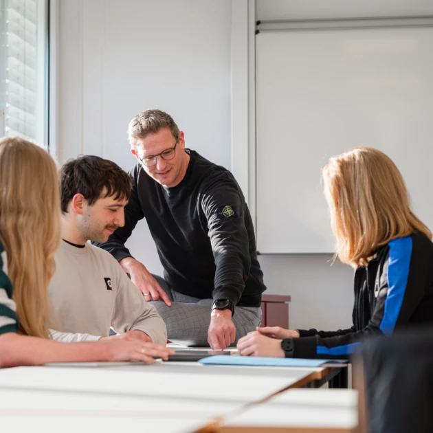 Gruppensituation in einem Seminarraum. 3 Studierende ( 2 Frauen, 1 Mann) sitzen einander an einem Tisch gegenüber und haben Lernmaterialien zwischen sich ausgbreitet. Sie tragen Alltagskleidung und blicken konzentriert auf die Materialien. Hinter ihnen sitzt der Dozent auf dem Tisch und zeigt mit dem Finger auf ein Dokument auf dem Tisch, Er trägt Alltagskleidung und eine Brille.