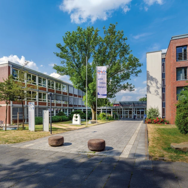 Haupteingang der EvH Bochum: Auf der rechten und linken Seite stehen große braun gepflasterte Hochschulgebäude mit vielen Fenstern. In der Mitte steht ein großer grüner Baum und eine Flagge mit dem rot-blauem Logo der Hochschule. Links und rechts neben den Gebäuden gibt es grüne Wiesen und einen Fahrradständer. Links vor der Gebäude steht eine weiße kleine Skulptur.